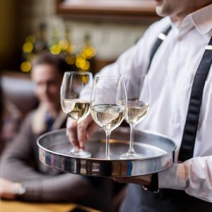 kelner w białej koszuli niesie 3 kieliszki białego wina na tacy